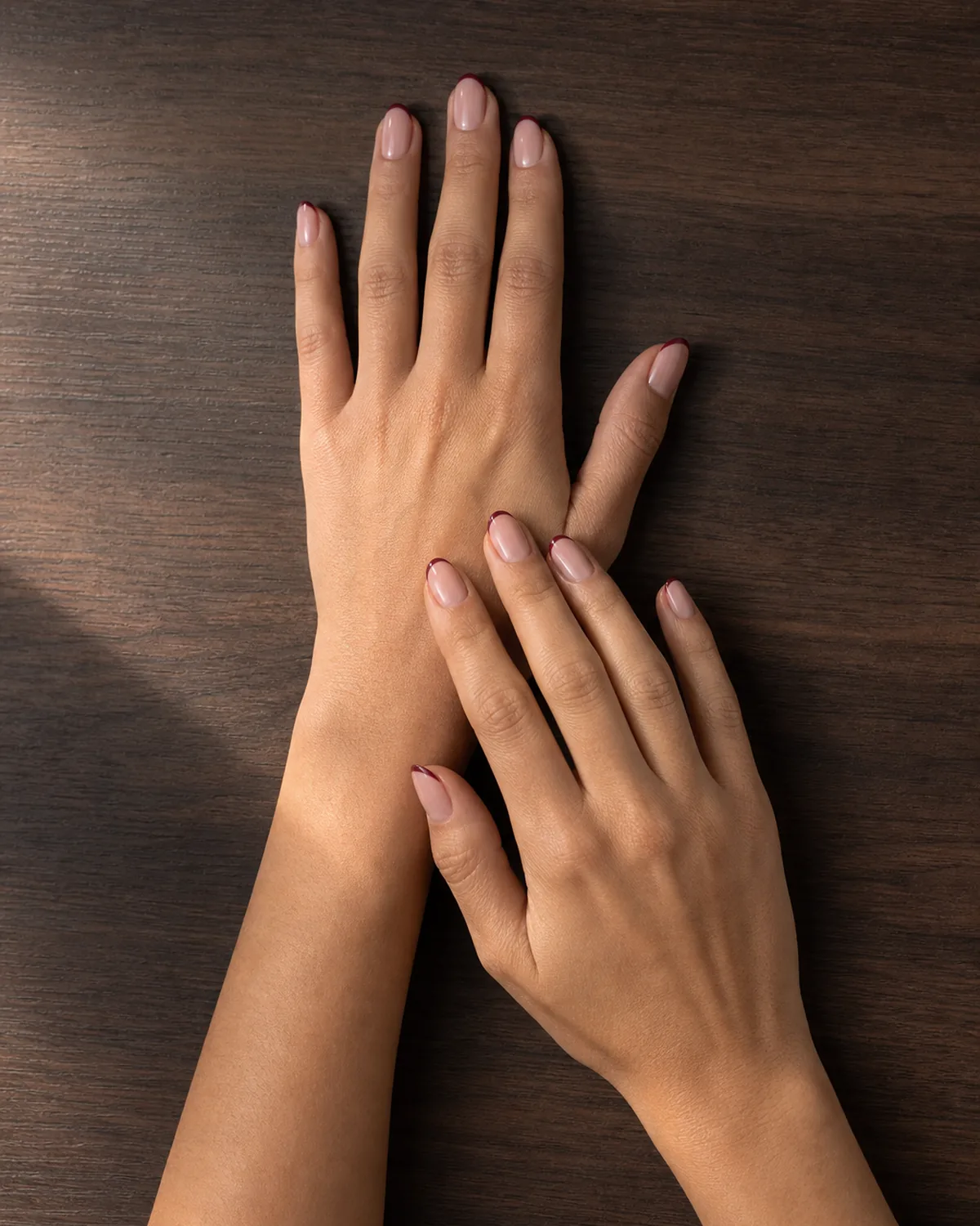 Two hands on dark wood showing thin burgundy tip lines on sheer pink base on oval nails