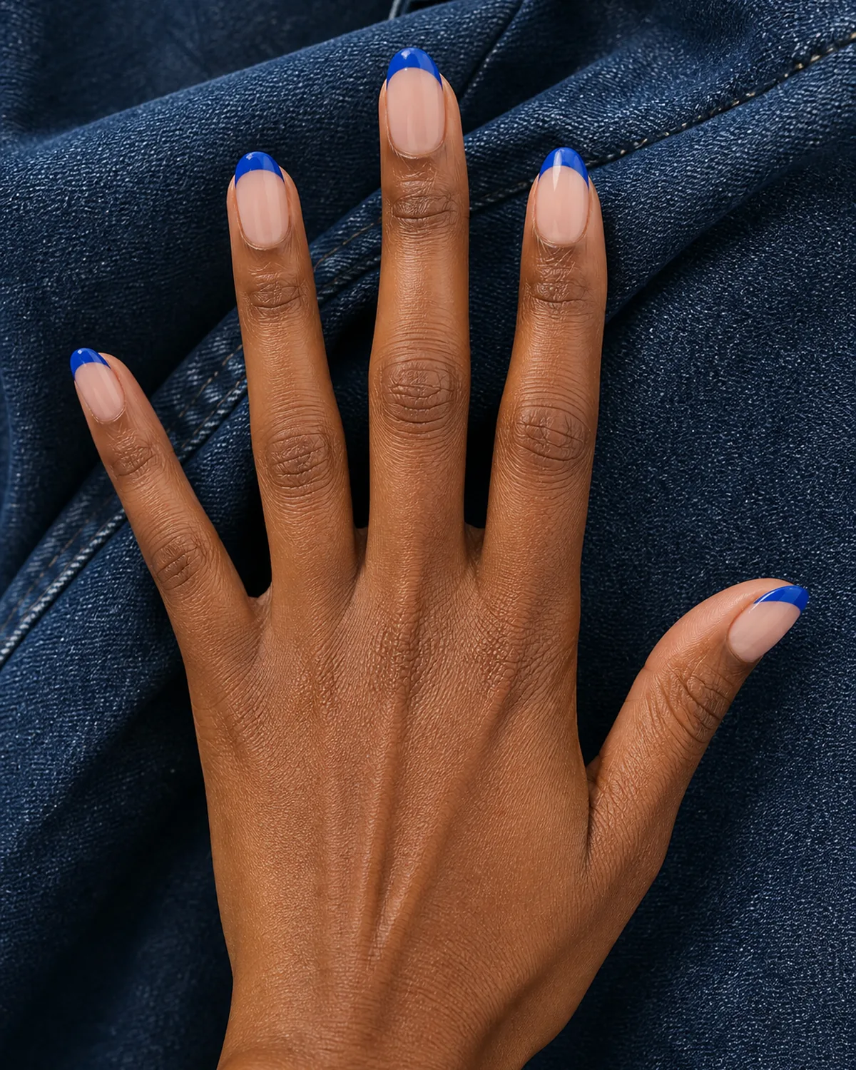 Single hand with cobalt blue colored french tips on medium oval nails against a folded dark denim jacket