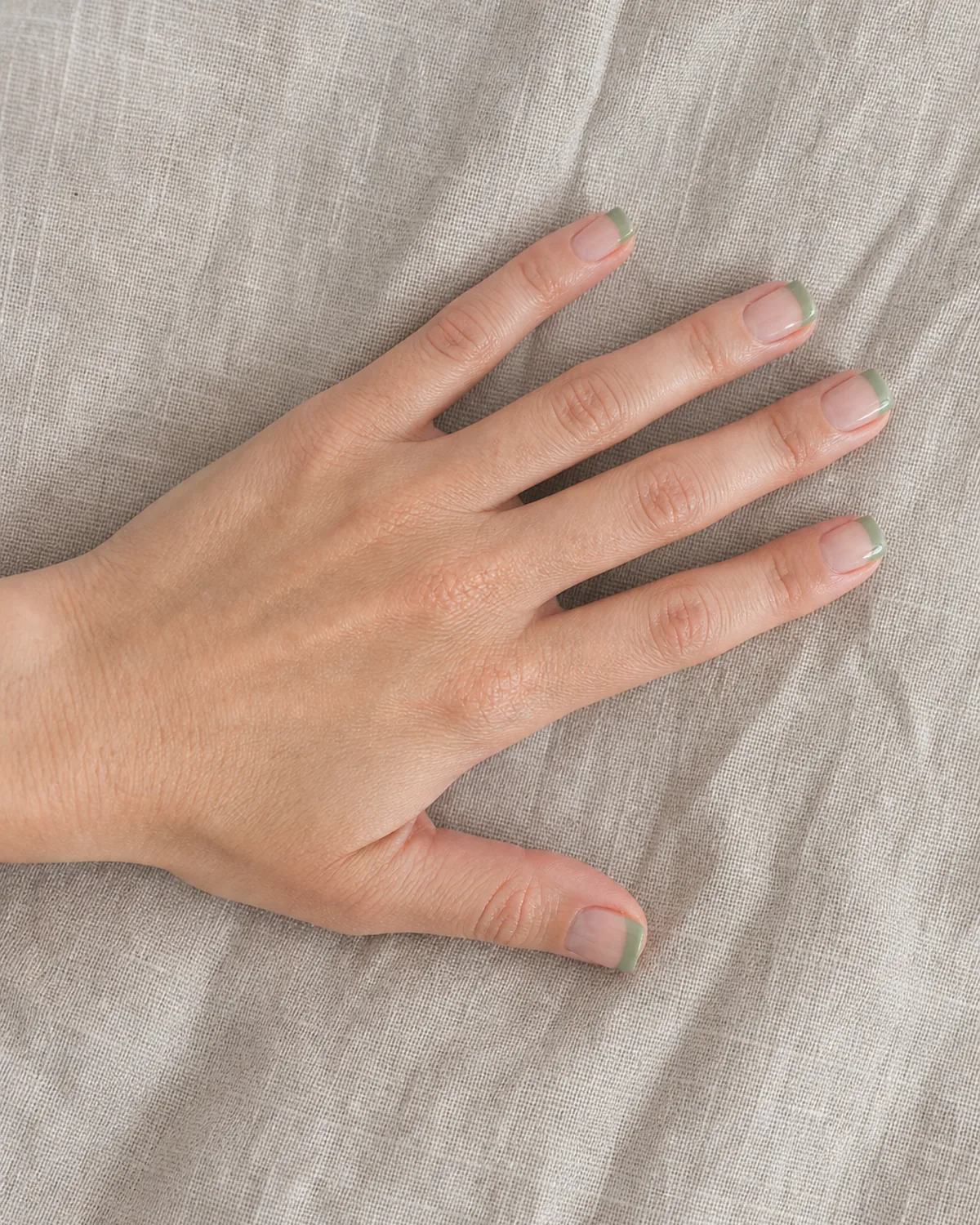 Single hand with sage green colored french tips on short square nails lying flat on a textured linen napkin