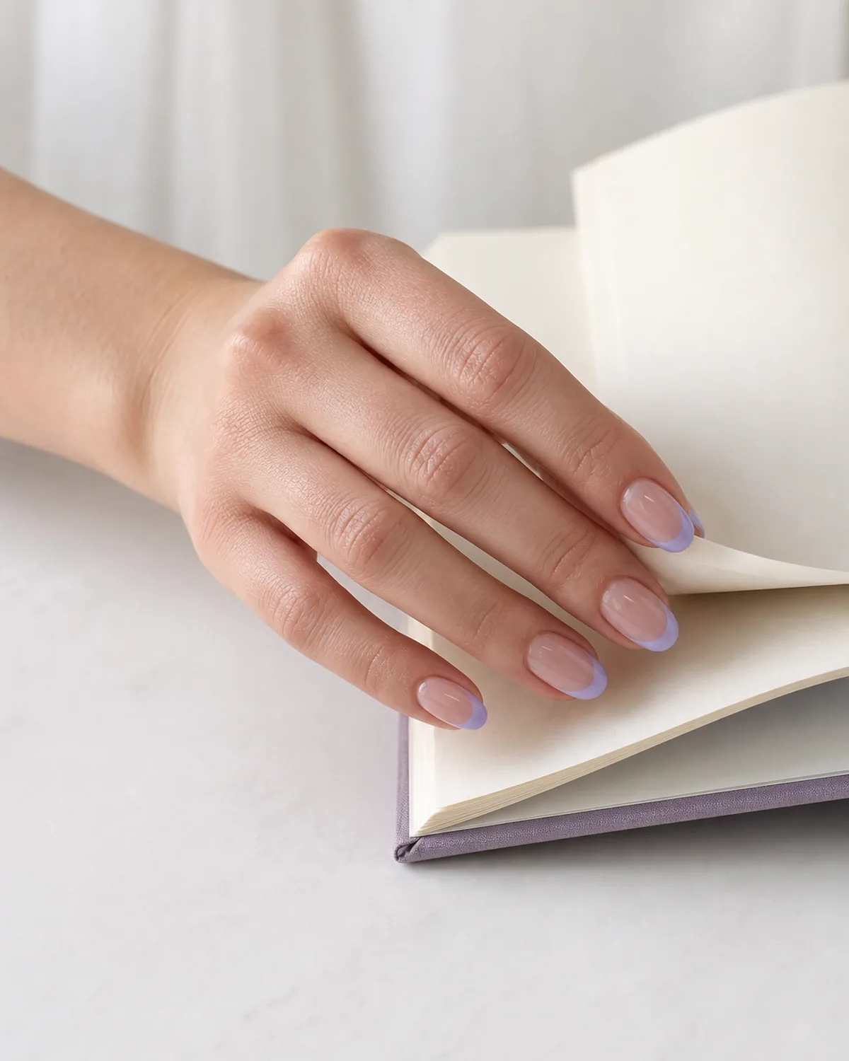 Single hand with soft lavender colored french tips on oval nails turning a page of an open book