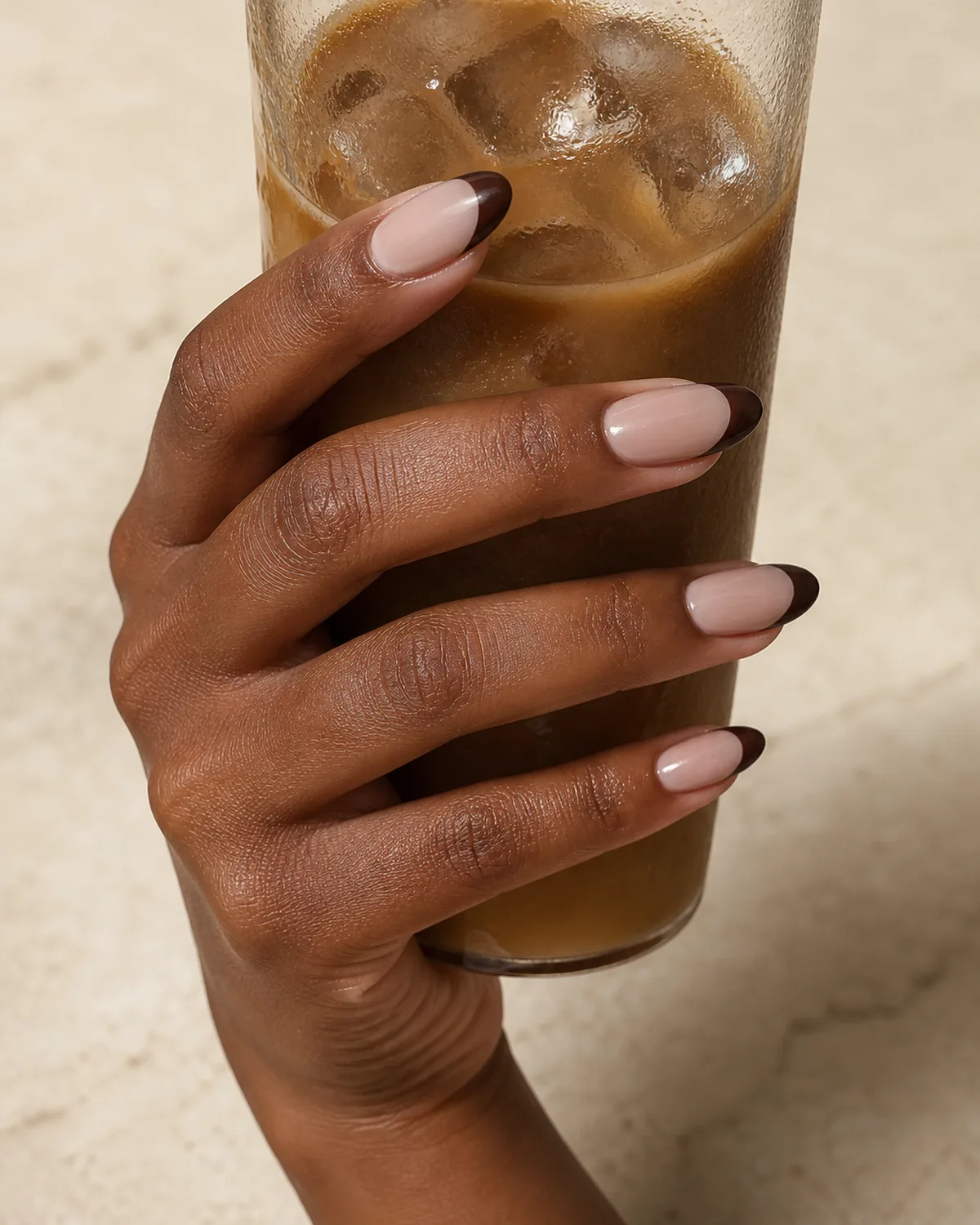 Single hand with deep chocolate brown colored french tips on almond nails wrapped around a tall iced coffee glass