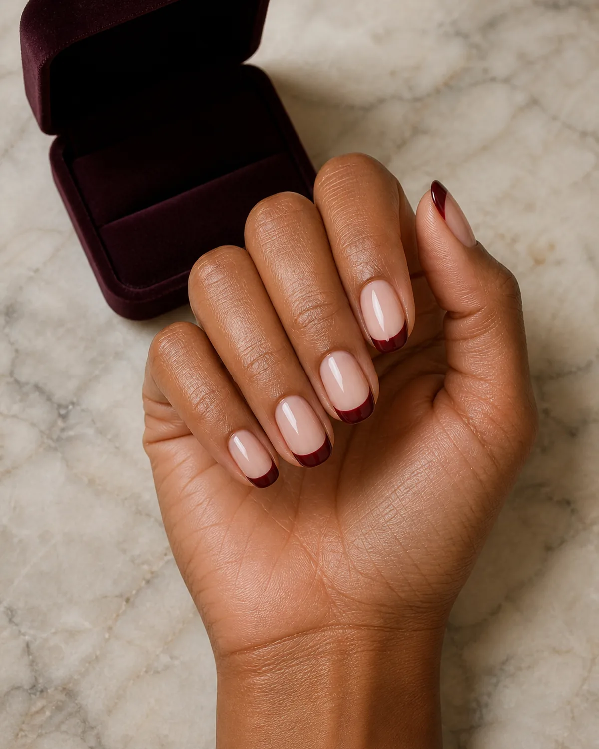 Single hand with deep burgundy colored french tips on medium round nails beside an open velvet jewelry box on marble