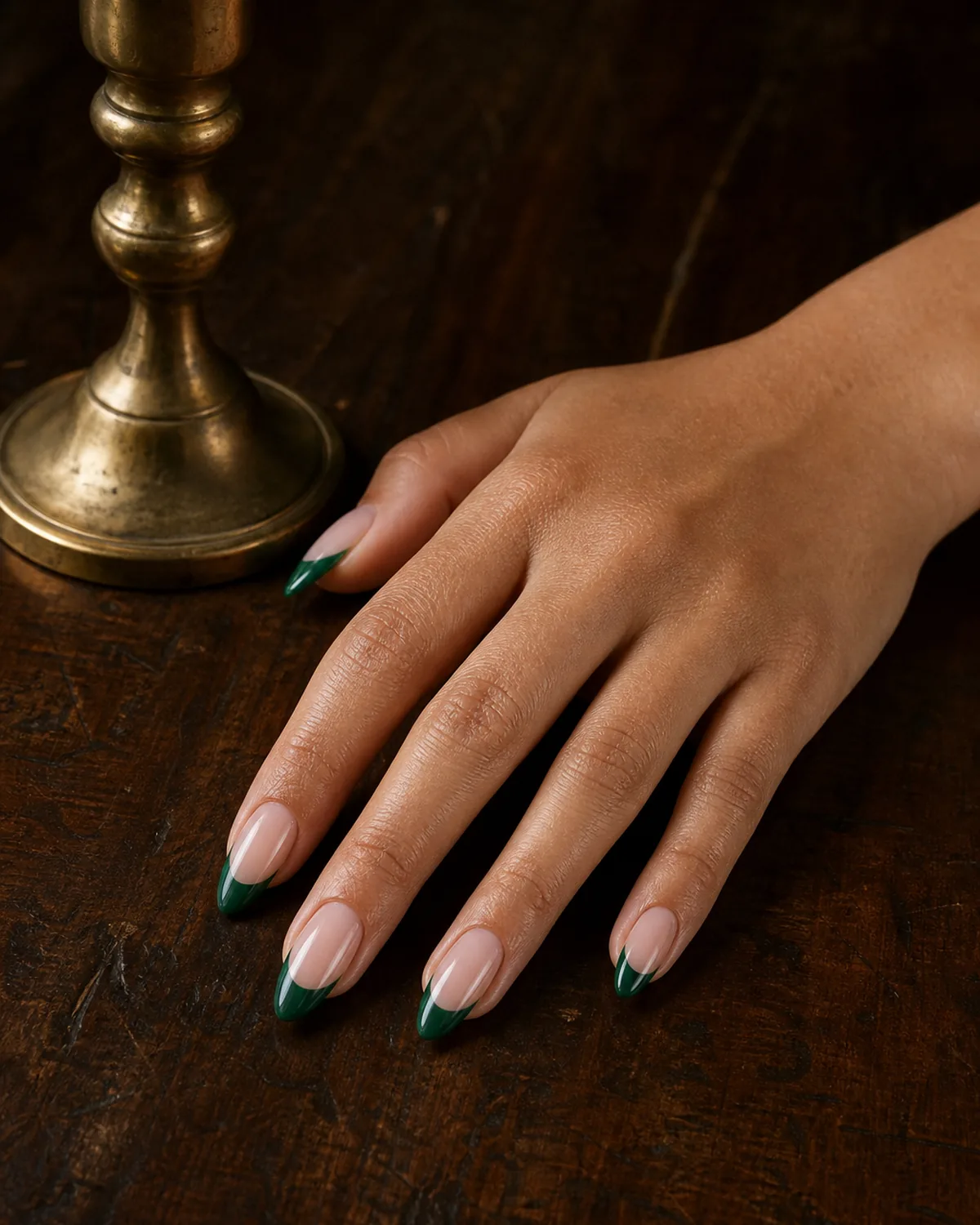 Single hand with rich emerald green colored french tips on almond nails resting on dark wood near a brass candlestick
