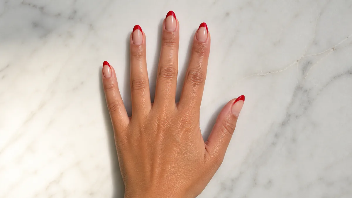 Single hand with cherry-red colored french tips on almond nails resting on white marble countertop