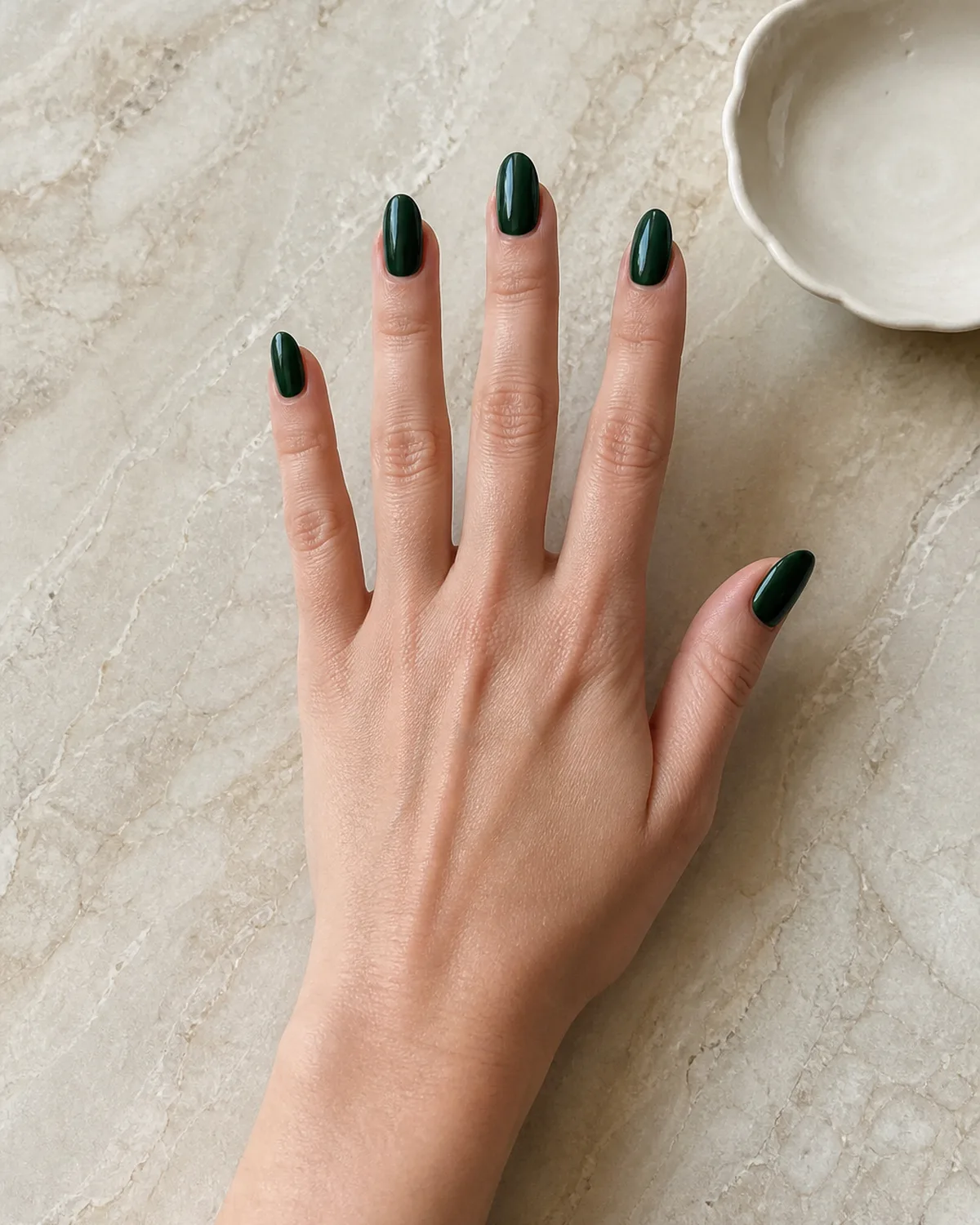 Single hand with deep forest green glossy crème nail polish on medium almond shaped nails resting palm-down on cream marble countertop beside small white ceramic dish