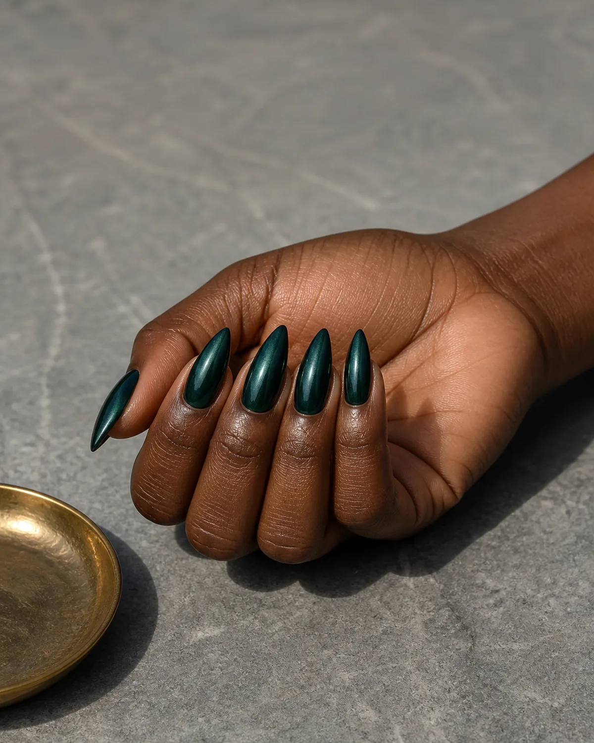 Single hand with dark pine green metallic chrome nail polish on medium stiletto shaped nails resting on its side on gray stone surface beside small brass dish