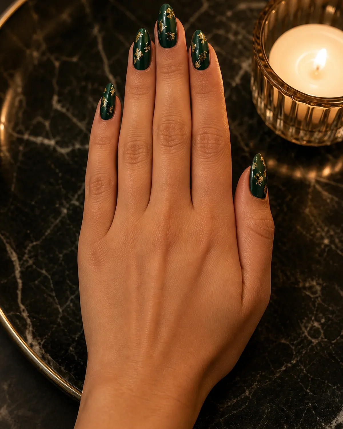 Single hand with deep British racing green nail polish with scattered irregular gold foil flecks on almond shaped nails beside gold-rimmed candle on dark marble tray