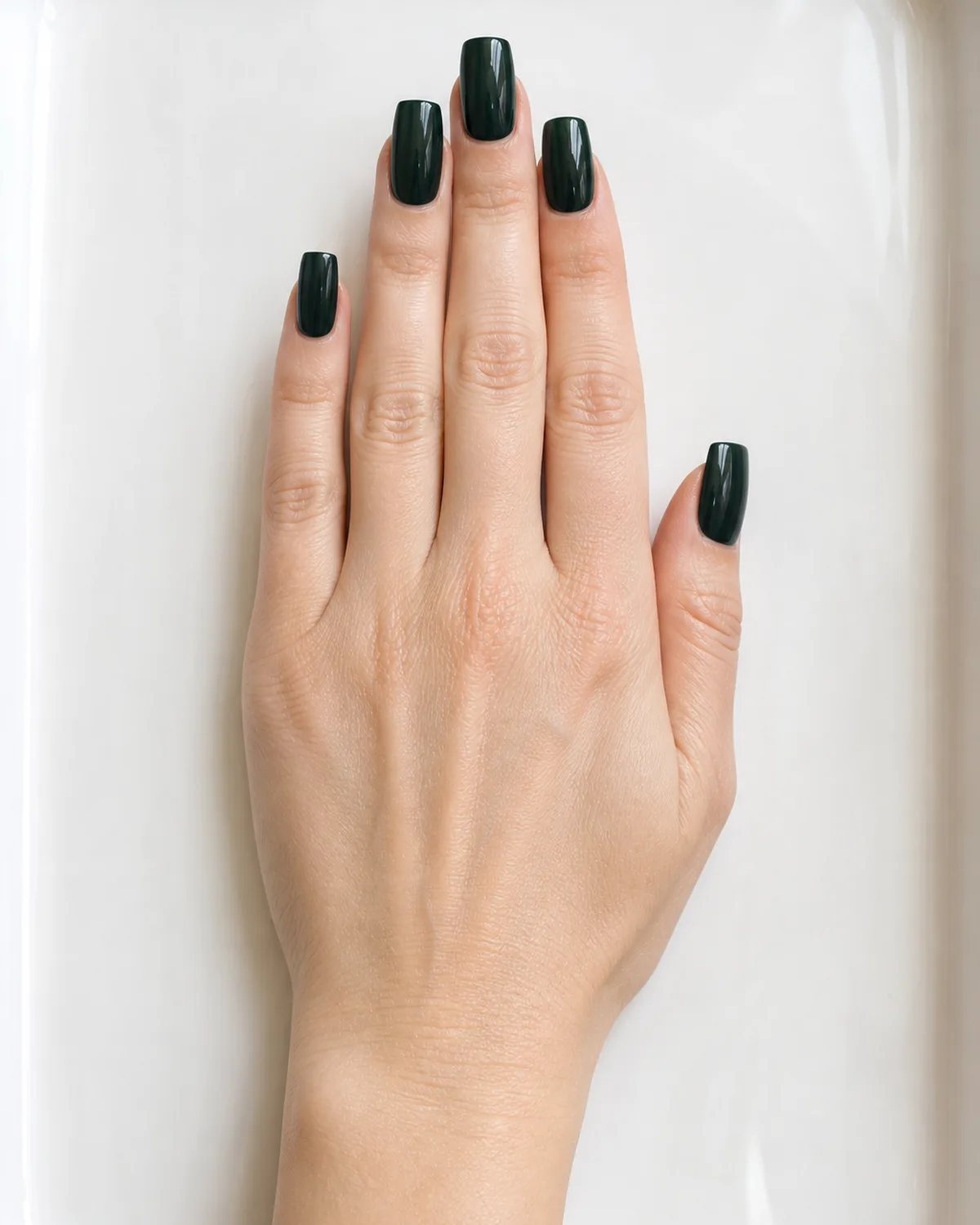 Single hand with deep Brunswick green blue-leaning dark green crème nail polish on long square shaped nails resting flat on white lacquered tray