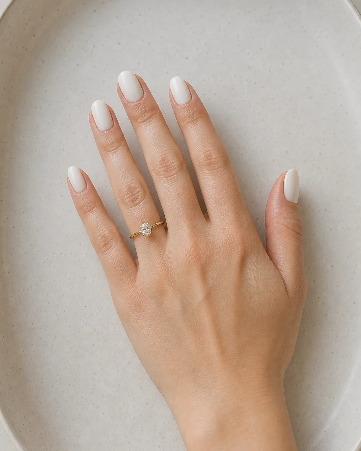 Single left hand with opaque milky white gel polish on medium oval nails resting palm down on ivory ceramic tray with engagement ring on ring finger