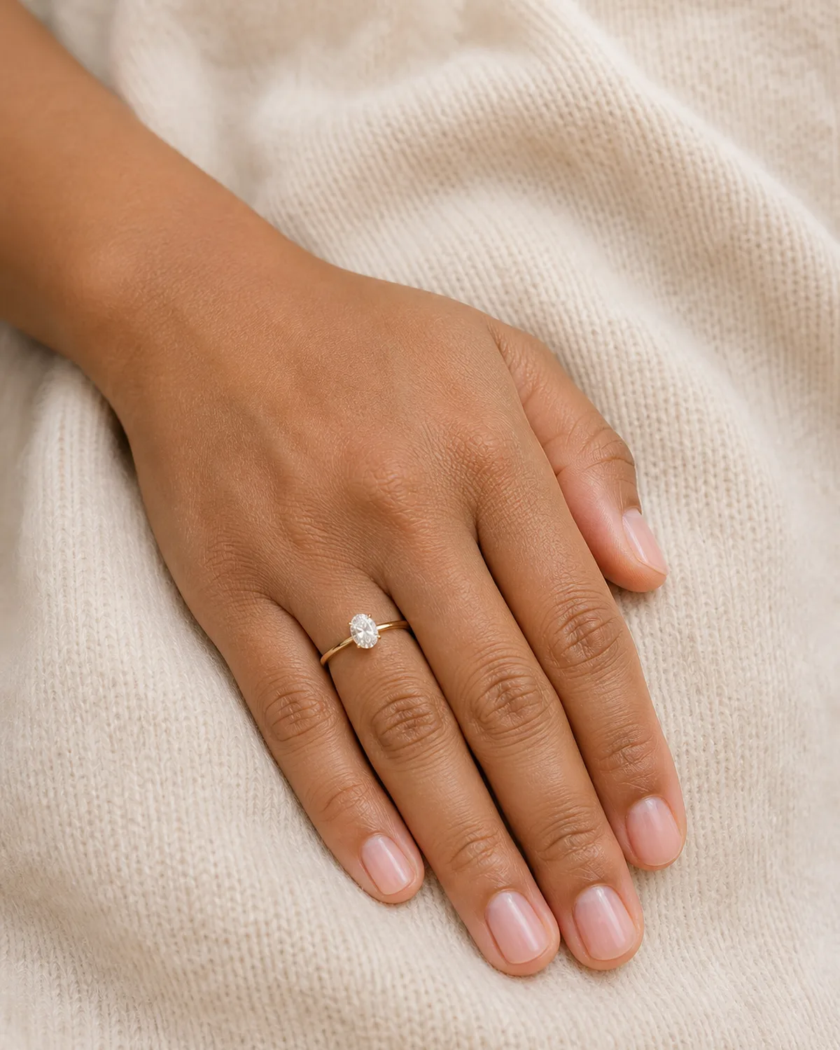 Single left hand with one-coat sheer pink polish on short round nails resting on cream cashmere sweater surface with engagement ring on ring finger