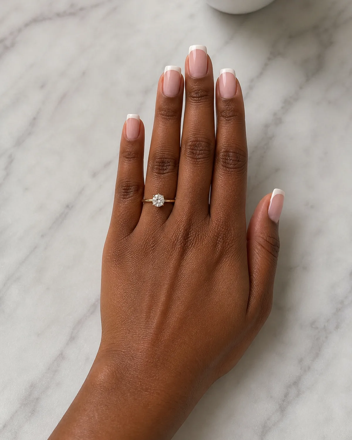 Single left hand with classic white French tips on medium square nails palm down on pale gray marble surface near white coffee cup with engagement ring visible