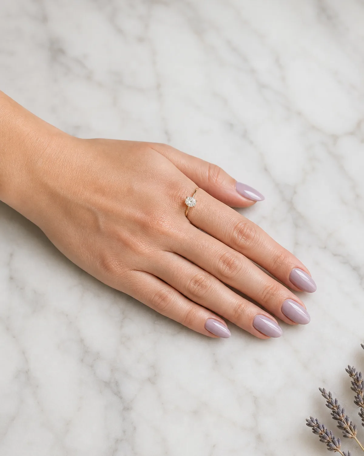 Single left hand with muted dusty lavender cream polish on medium almond nails on white marble surface with dried lavender sprig at frame edge and engagement ring on ring finger