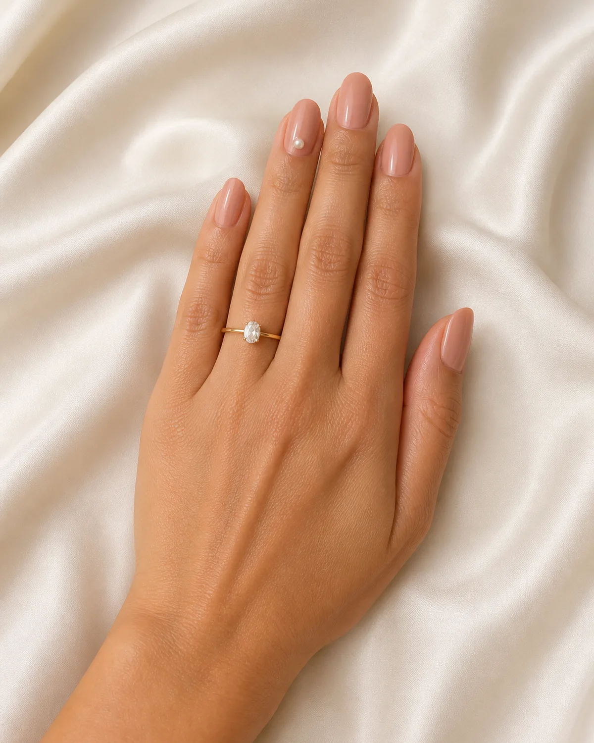 Single left hand with warm nude polish on medium oval nails and a small faux pearl stud on ring finger nail on cream satin fabric with engagement ring visible
