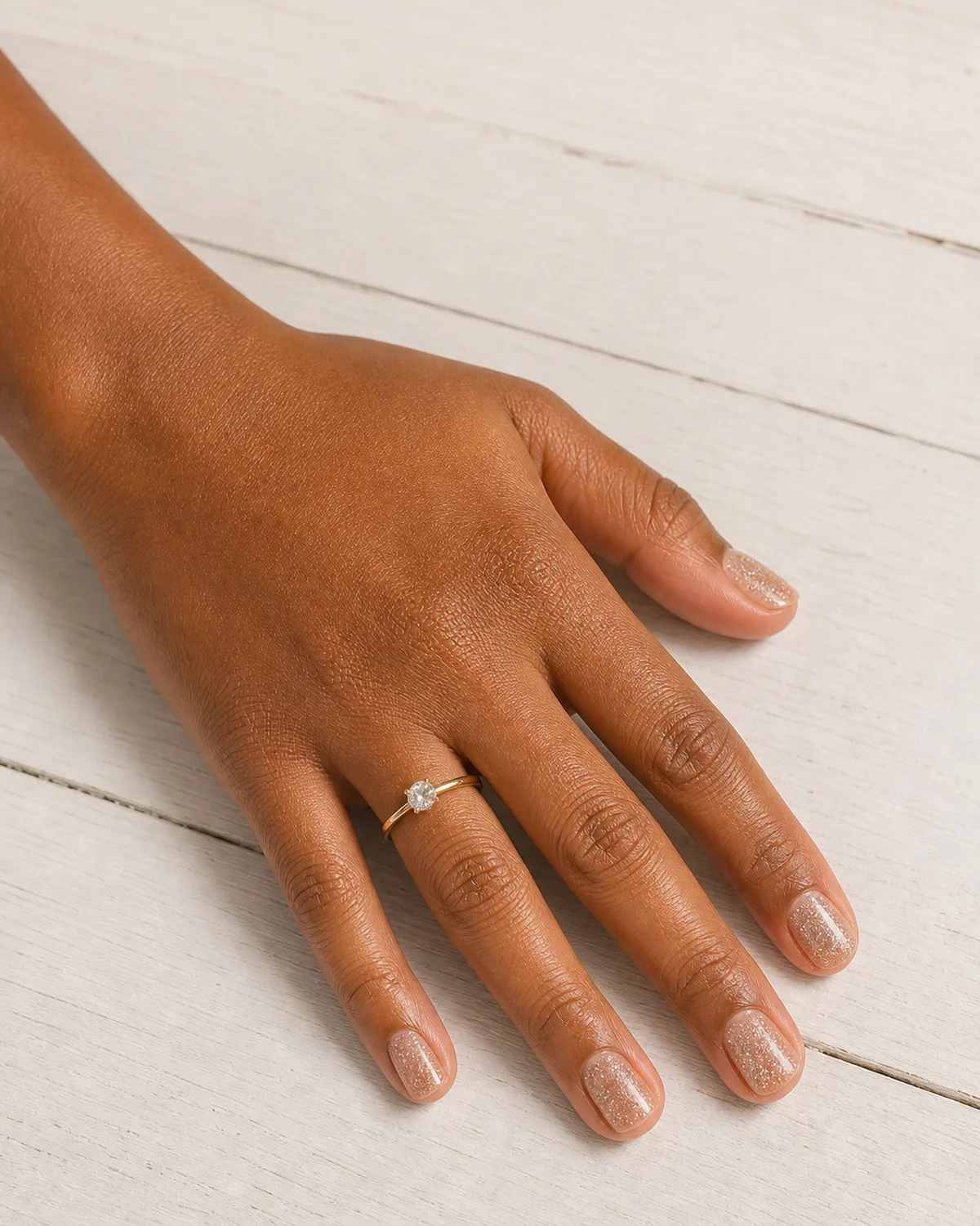 Single left hand with fine champagne gold micro glitter in clear base on short square nails resting on white painted wood surface with engagement ring on ring finger