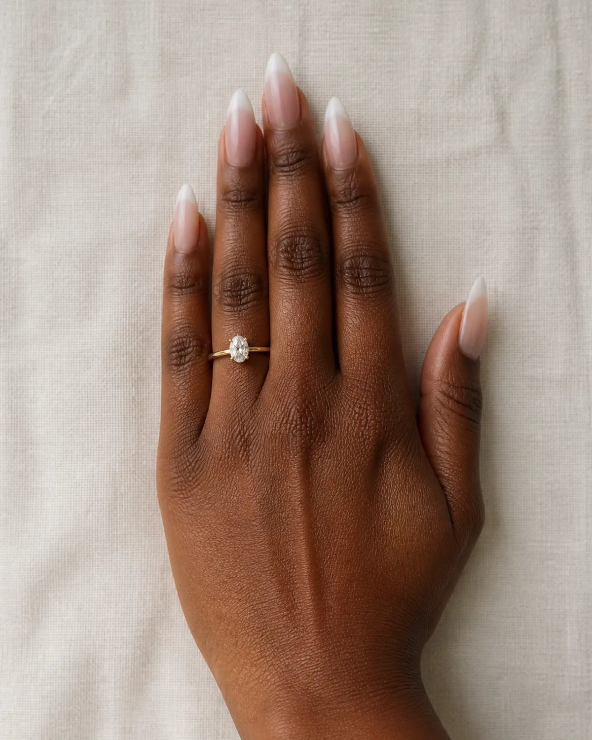 Single left hand with nude-to-white gradient ombre on medium stiletto nails resting on off-white linen tablecloth with engagement ring on ring finger