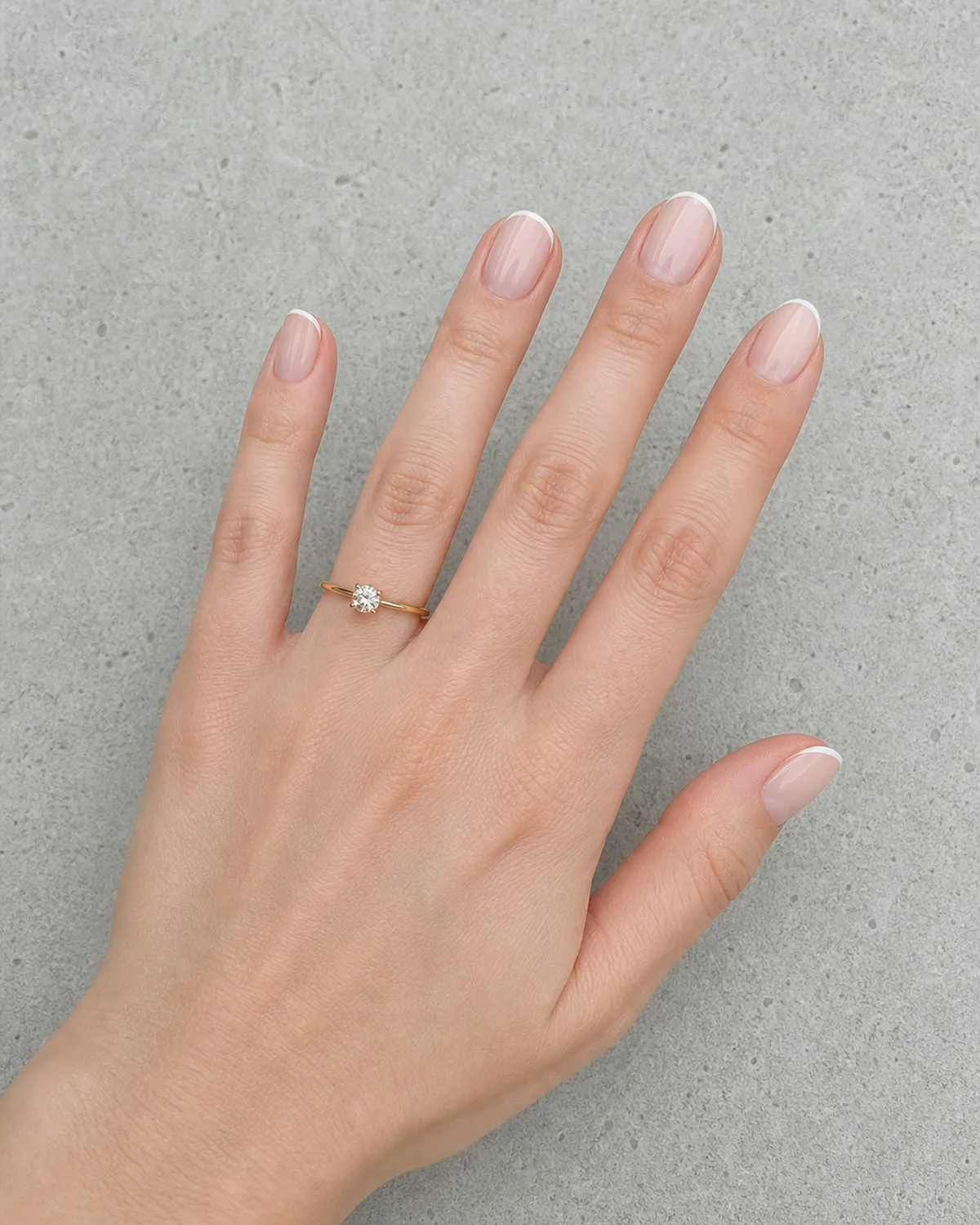 Single left hand with thin white micro French line at free edge on medium oval nails on light gray concrete surface with engagement ring on ring finger