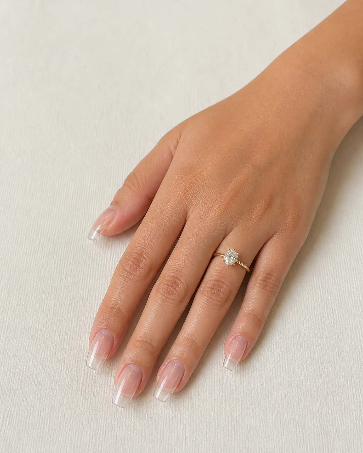 Single left hand with crystal clear transparent hard gel on medium coffin nails on cream textured paper surface with engagement ring on ring finger