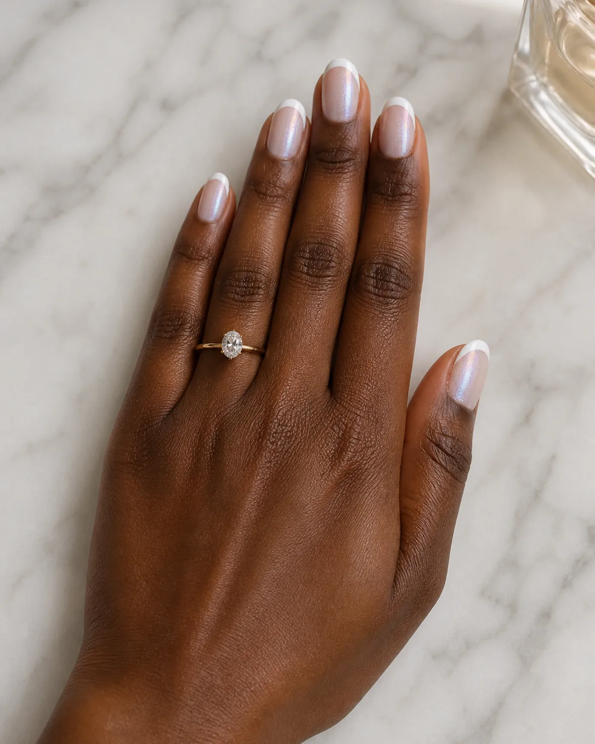 Single left hand with sheer shimmer iridescent base coat and crisp white French tips on medium oval nails on white marble surface near glass perfume bottle with engagement ring on ring finger