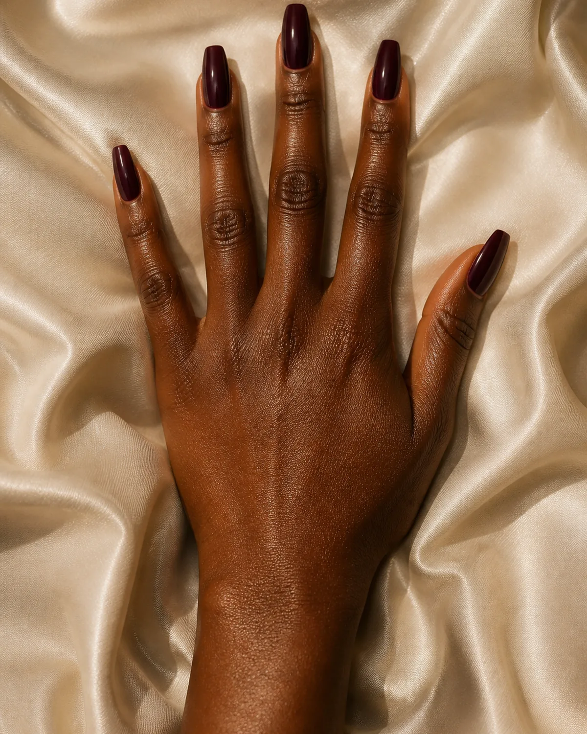 Single hand with rich aubergine plum gloss polish on medium-long coffin nails palm down on ivory satin fabric