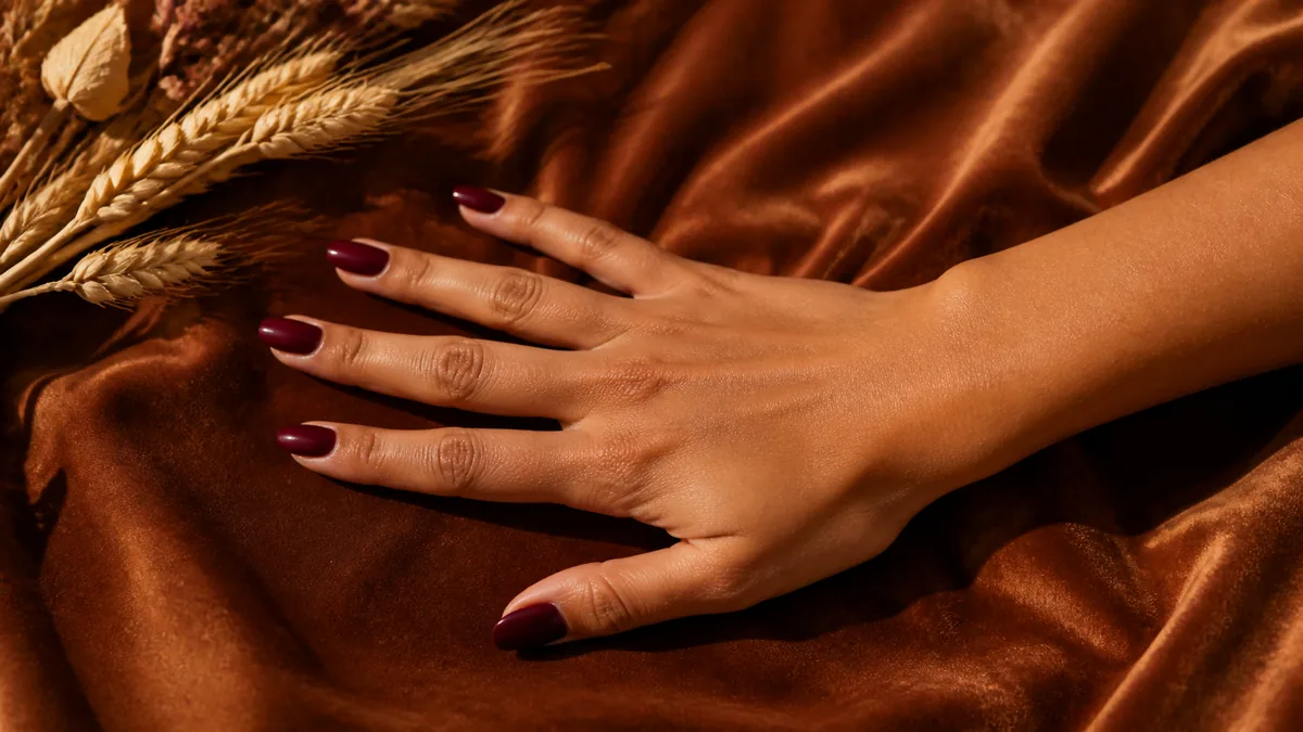 Single hand with deep burgundy gloss polish on medium almond nails palm down on rust-colored velvet fabric beside dried wheat stalks