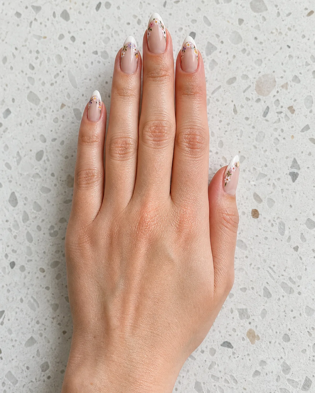 Single hand with nude nails with thin white french tip and tiny hand-painted wildflowers trailing from the smile line on long almond shape on white terrazzo