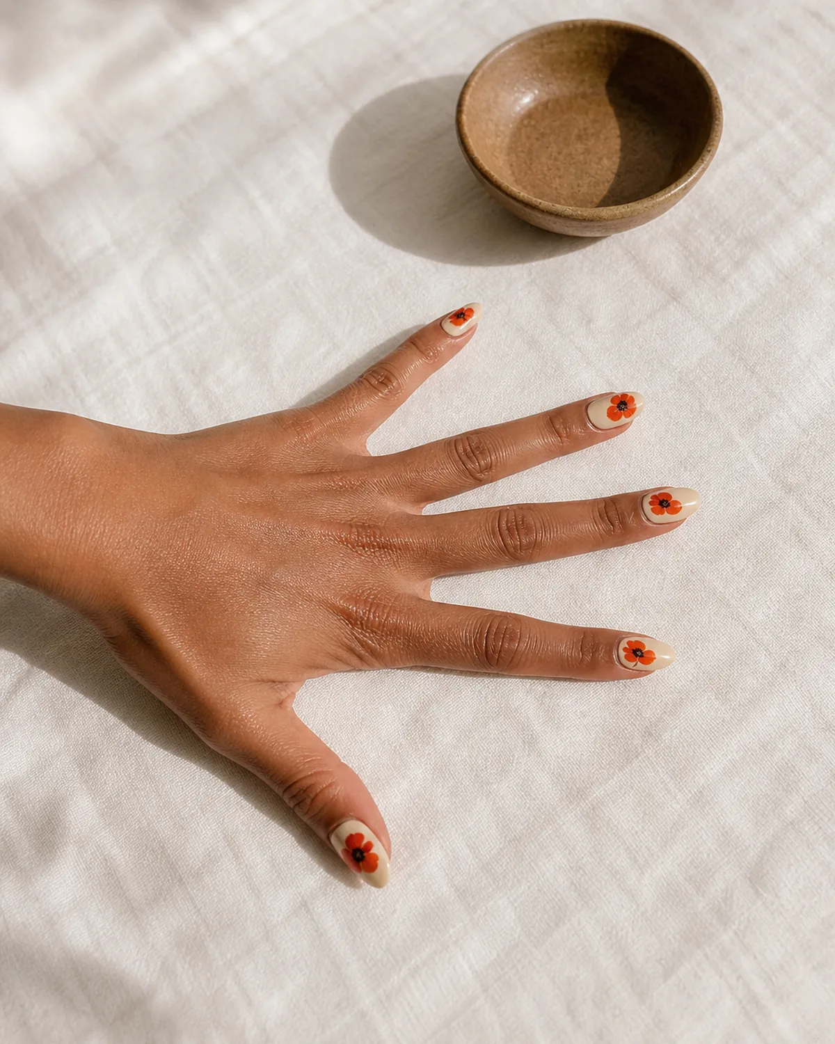 Single hand with vivid red-orange poppy nail art with dark centers on warm cream base medium almond nails wide splay on white linen beside brown ceramic bowl