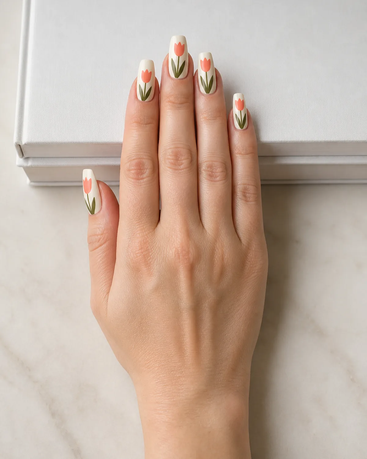 Single hand with graphic coral and green tulip silhouette nail art on creamy off-white base coffin nails resting flat on stack of white hardcover books