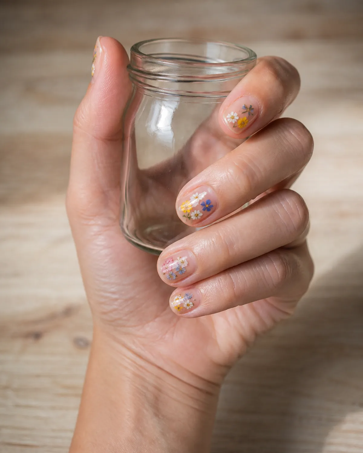 Single hand with tiny multi-colored wildflower garden nail art on clear glossy base short oval nails wrapped around small glass jar on pale wood desk