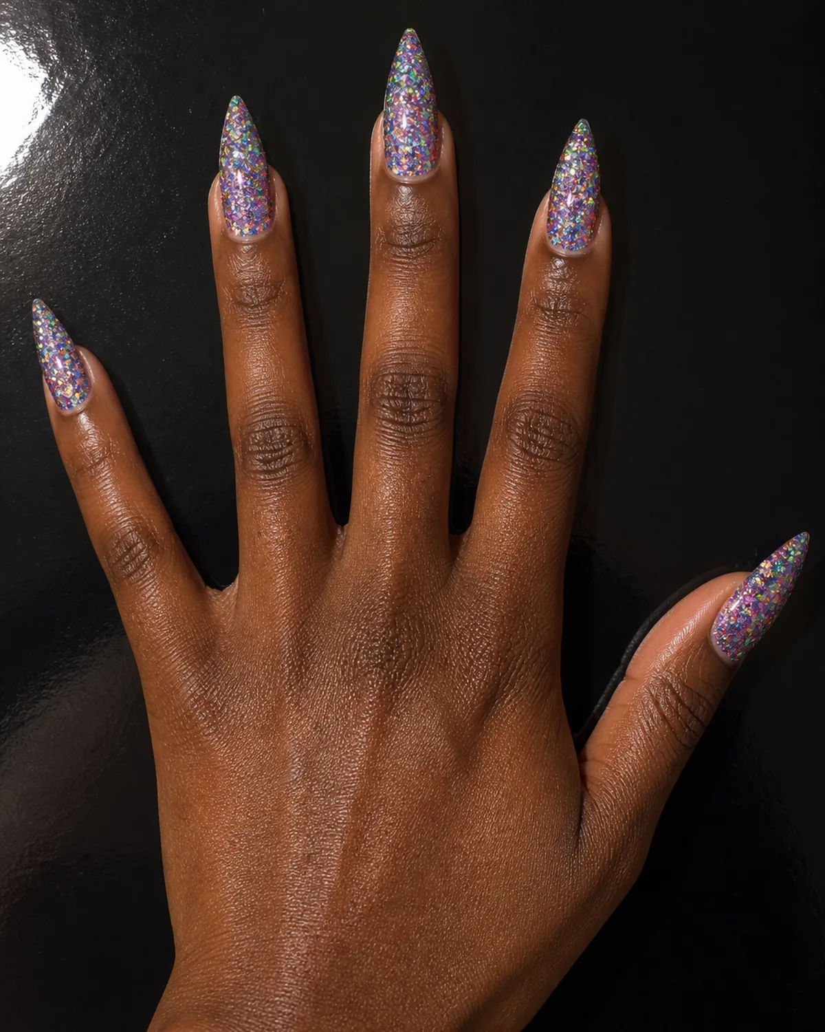 Single hand with holographic rainbow-shift glitter nails on stiletto shape with fingers fanned on a glossy black surface