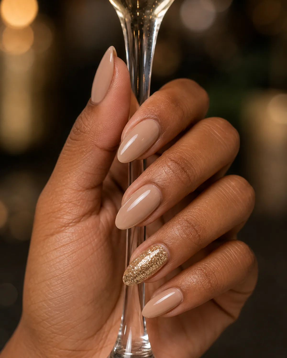Single hand with nude almond nails and one ring-finger accent nail covered in champagne gold glitter pinching a champagne flute stem