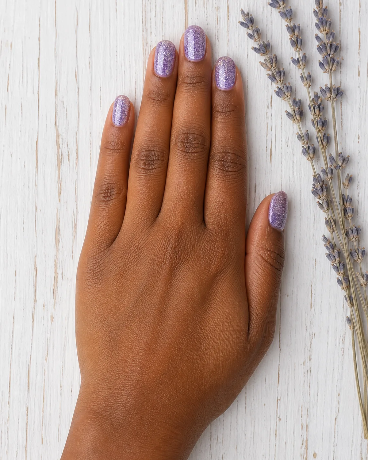 Single hand with lavender purple glitter nails on oval shape lying flat beside a sprig of dried lavender on white-painted wood