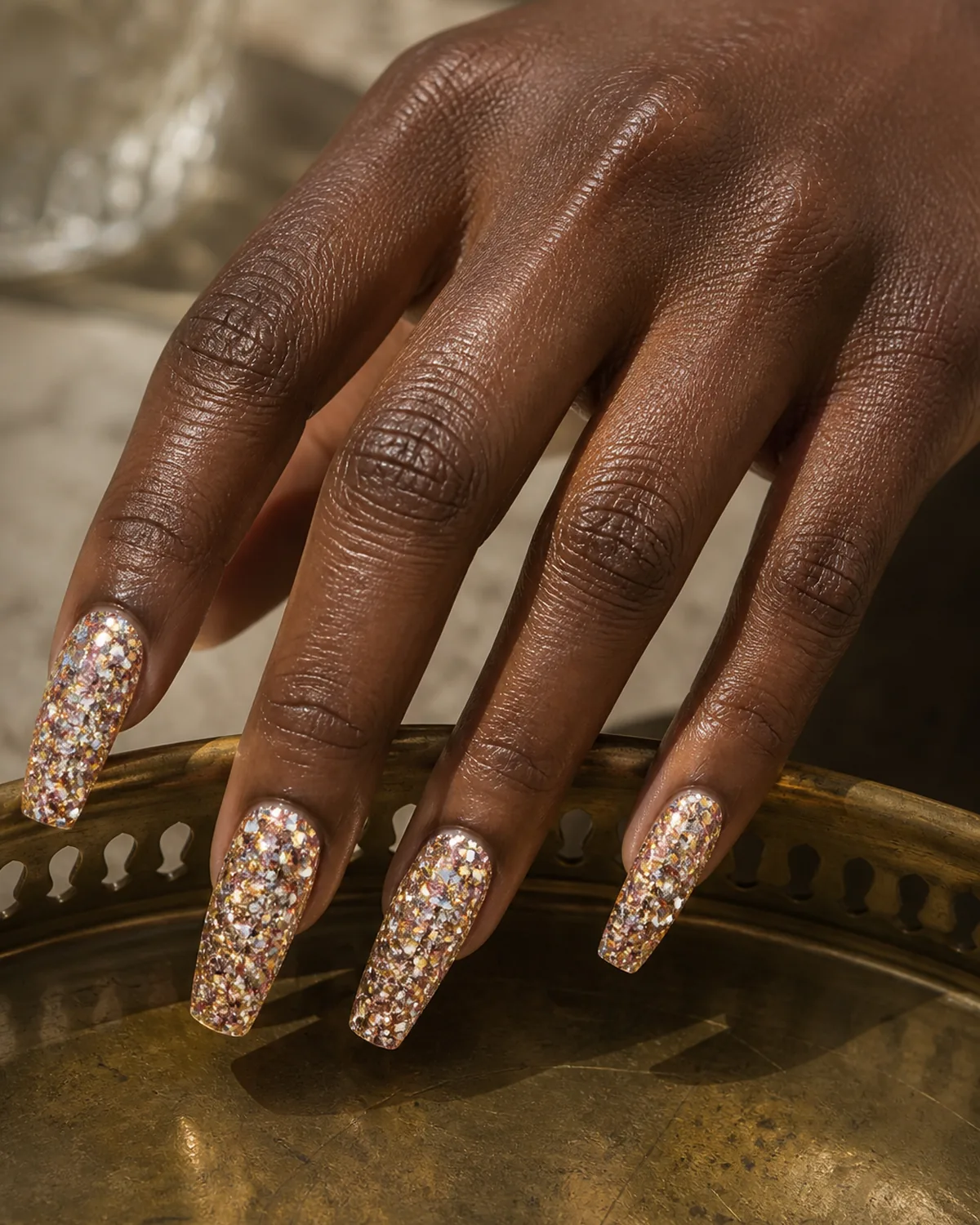 Single hand with mixed-metal glitter nails on coffin shape showing gold silver and rose gold particles combined with fingers extended along a brass tray edge