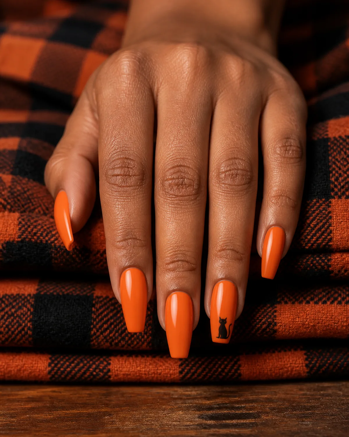 Single hand with bright pumpkin orange crème nail polish on coffin shaped nails with tiny black cat silhouette on ring finger resting on folded orange and black plaid flannel on wood surface