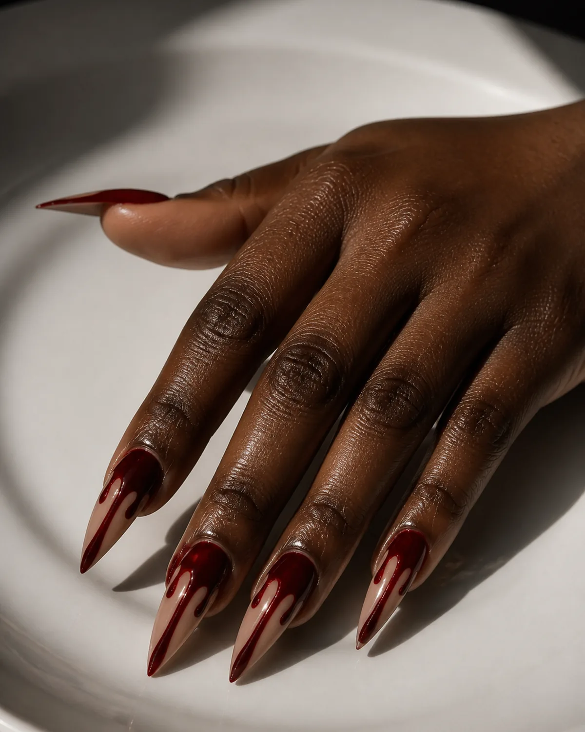 Single hand with sheer nude base on long stiletto shaped nails with glossy red polish dripping down from cuticle area mimicking blood drip effect resting on white ceramic plate in dramatic side-lighting