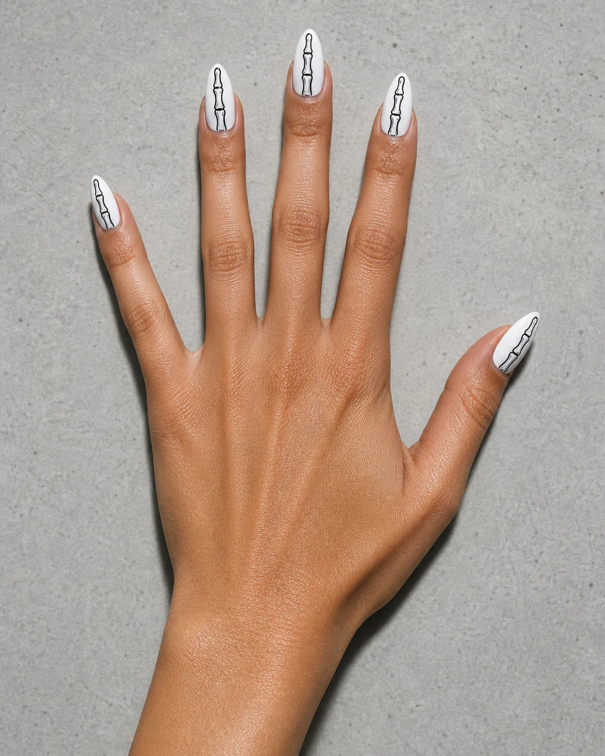 Single hand with crisp white nail polish on almond shaped nails with fine black line-art of skeleton bones drawn along each nail resting palm-flat on light gray concrete surface with fingers spread