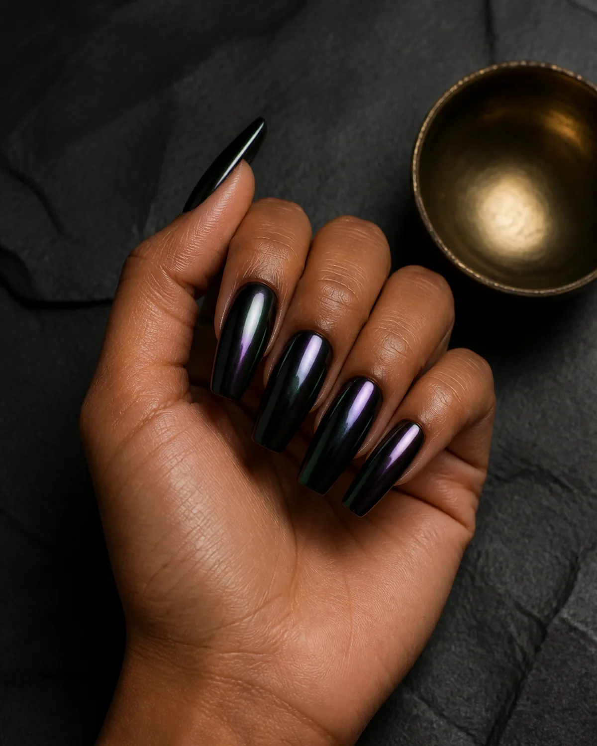 Single hand with jet black chrome nail polish that shifts to dark green and purple on long coffin shaped nails resting on dark stone surface beside small brass bowl