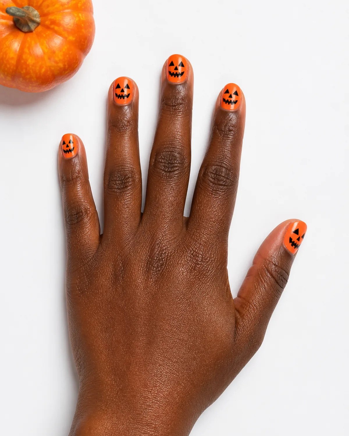 Single hand with bright orange nail polish on short round nails with simple carved pumpkin face designs in black on each nail resting palm-down on white surface beside small orange gourd
