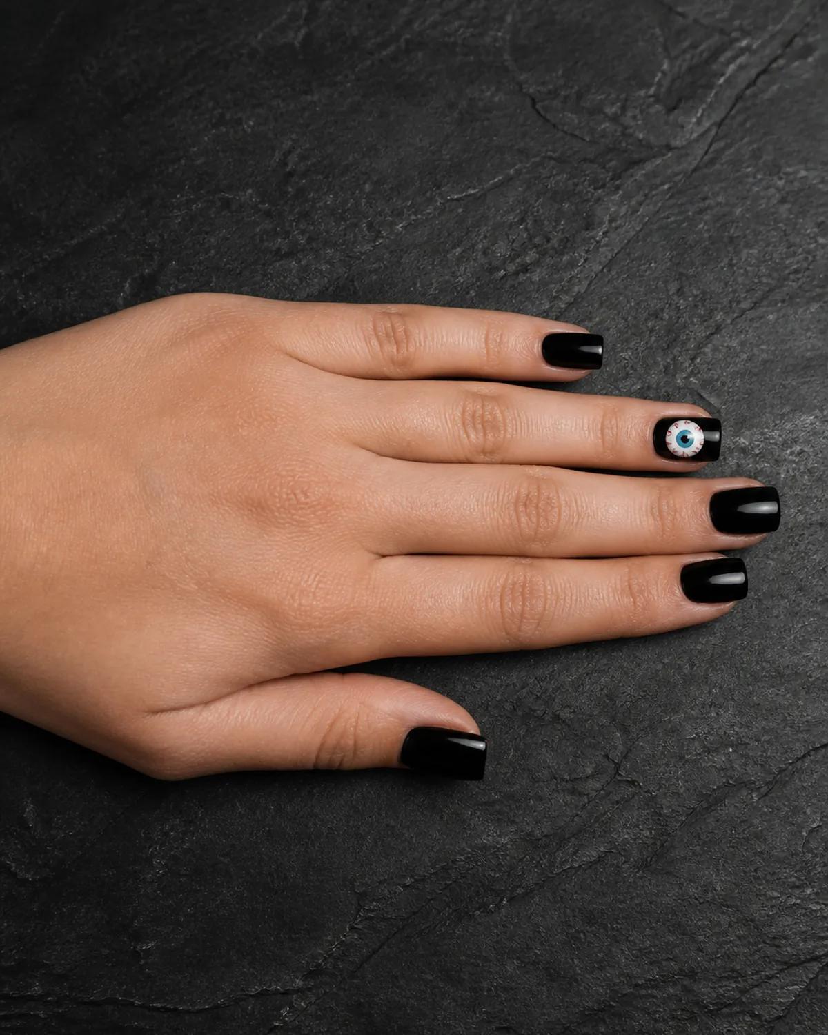 Single hand with glossy black nail polish on square nails with one accent nail featuring a hand-painted eyeball design with white circle colored iris and black pupil on dark charcoal slate surface