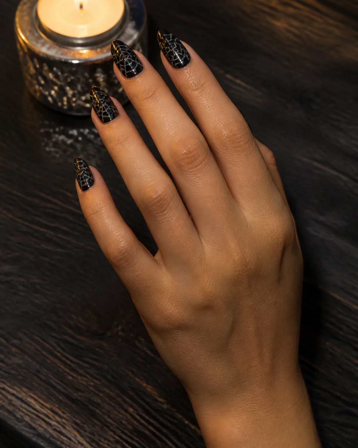 Single hand with glossy black nail polish on almond nails with fine silver glitter cobweb pattern stamped across each nail resting on dark wood table beside small silver tealight holder
