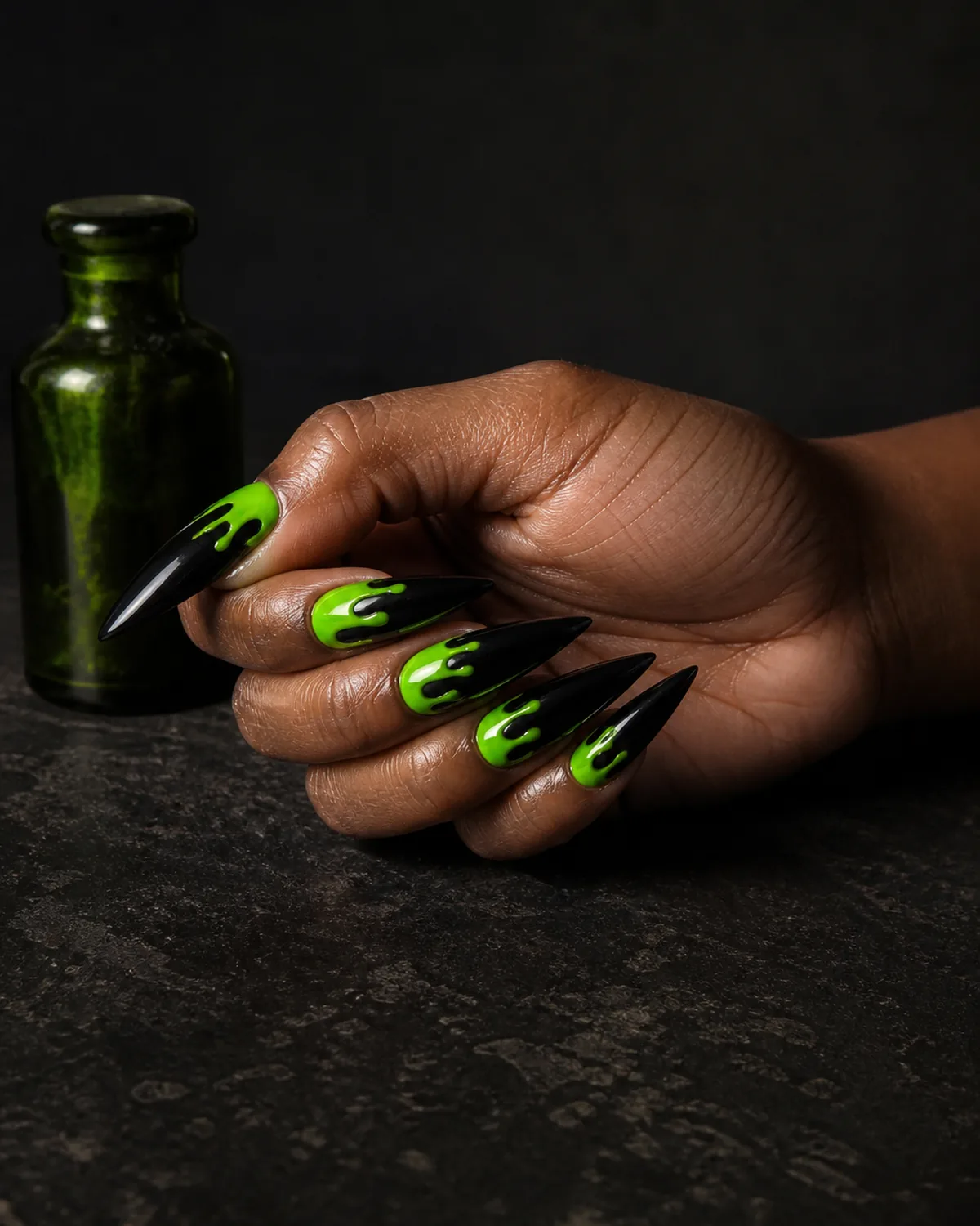 Single hand with jet black nail polish on long stiletto nails with neon slime-green polish dripping from cuticle on each nail resting on its side on dark surface beside small green glass bottle