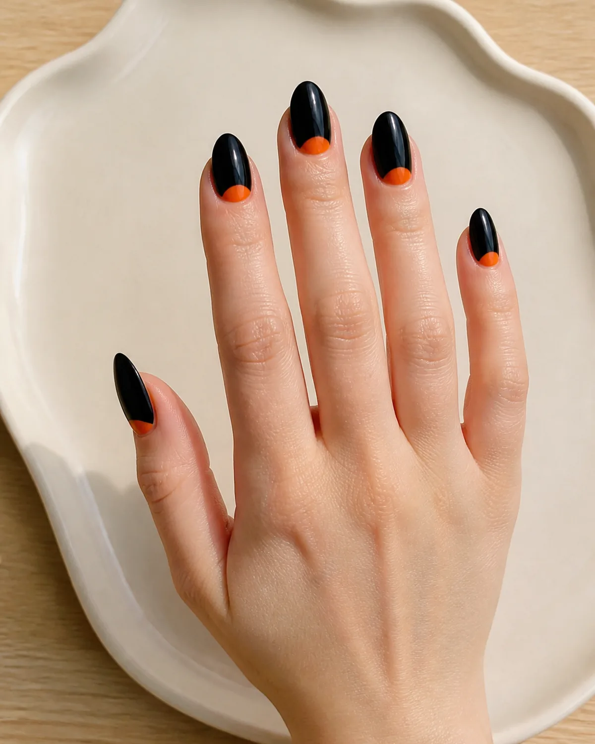 Single hand with black crème nail polish and bright orange half-moon at cuticle on medium almond nails resting on cream ceramic tray on light wood surface