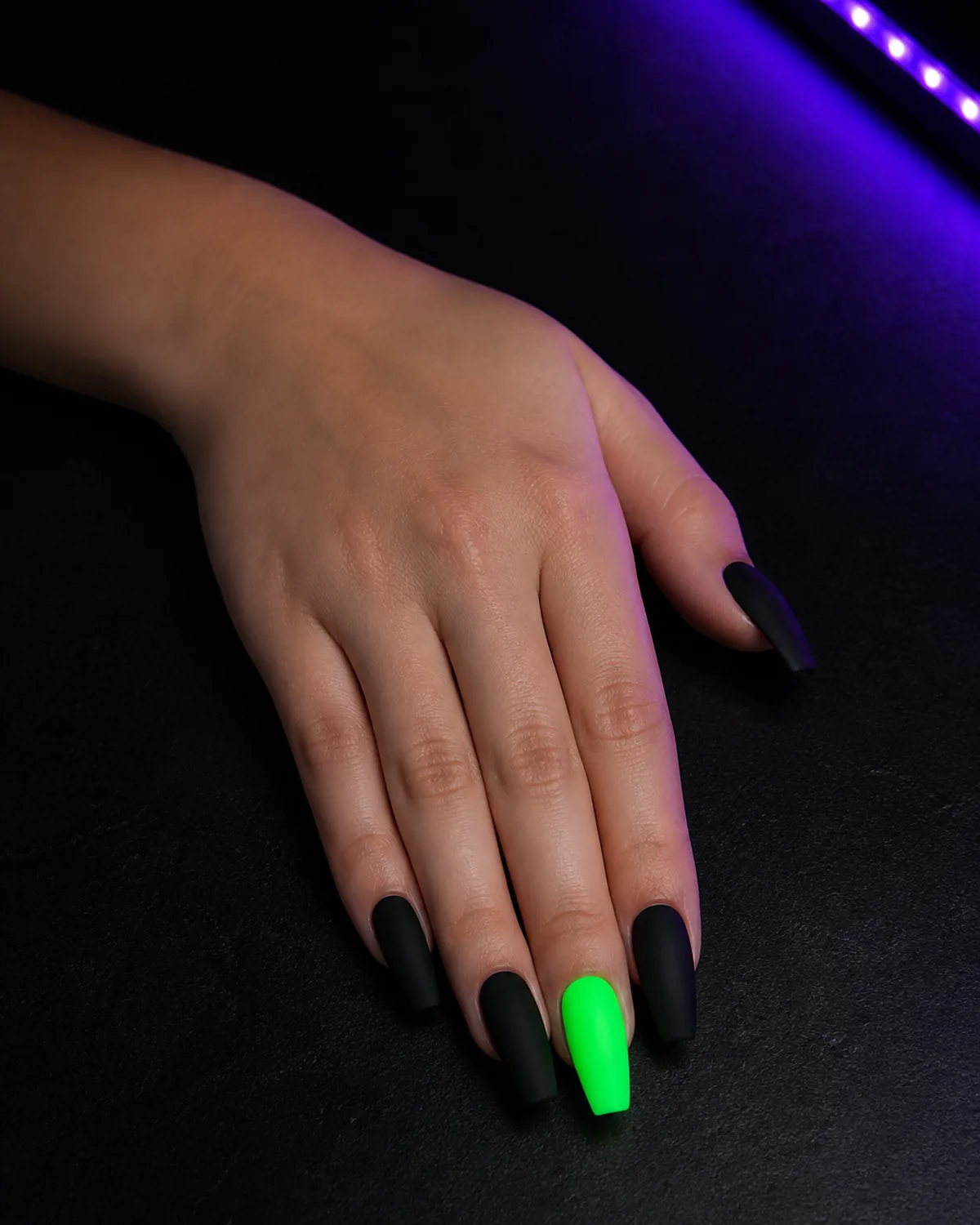 Single hand with matte black nail polish on coffin nails with one or two accent nails in glow-in-the-dark neon green polish resting palm-down on black surface with UV LED purple glow from one side