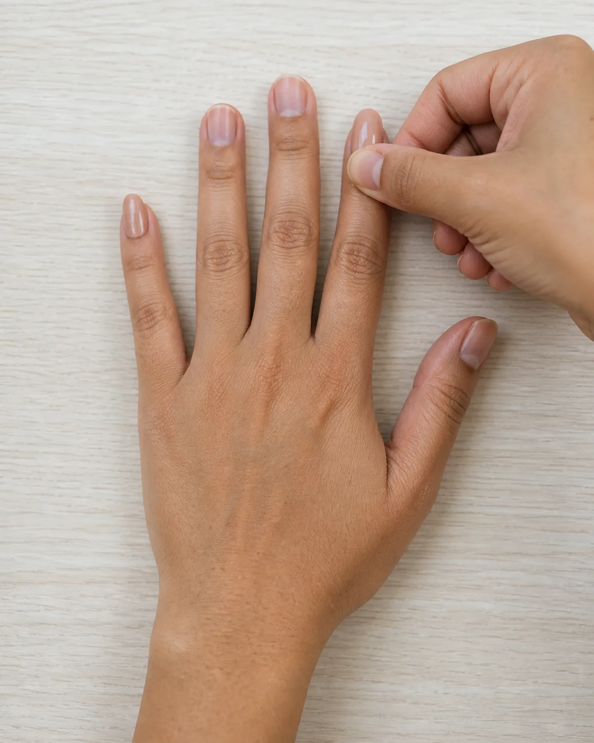 Single hand flat on pale wood desk with the thumb of other hand pressing down firmly on a just-applied glossy nude press-on nail on the index finger