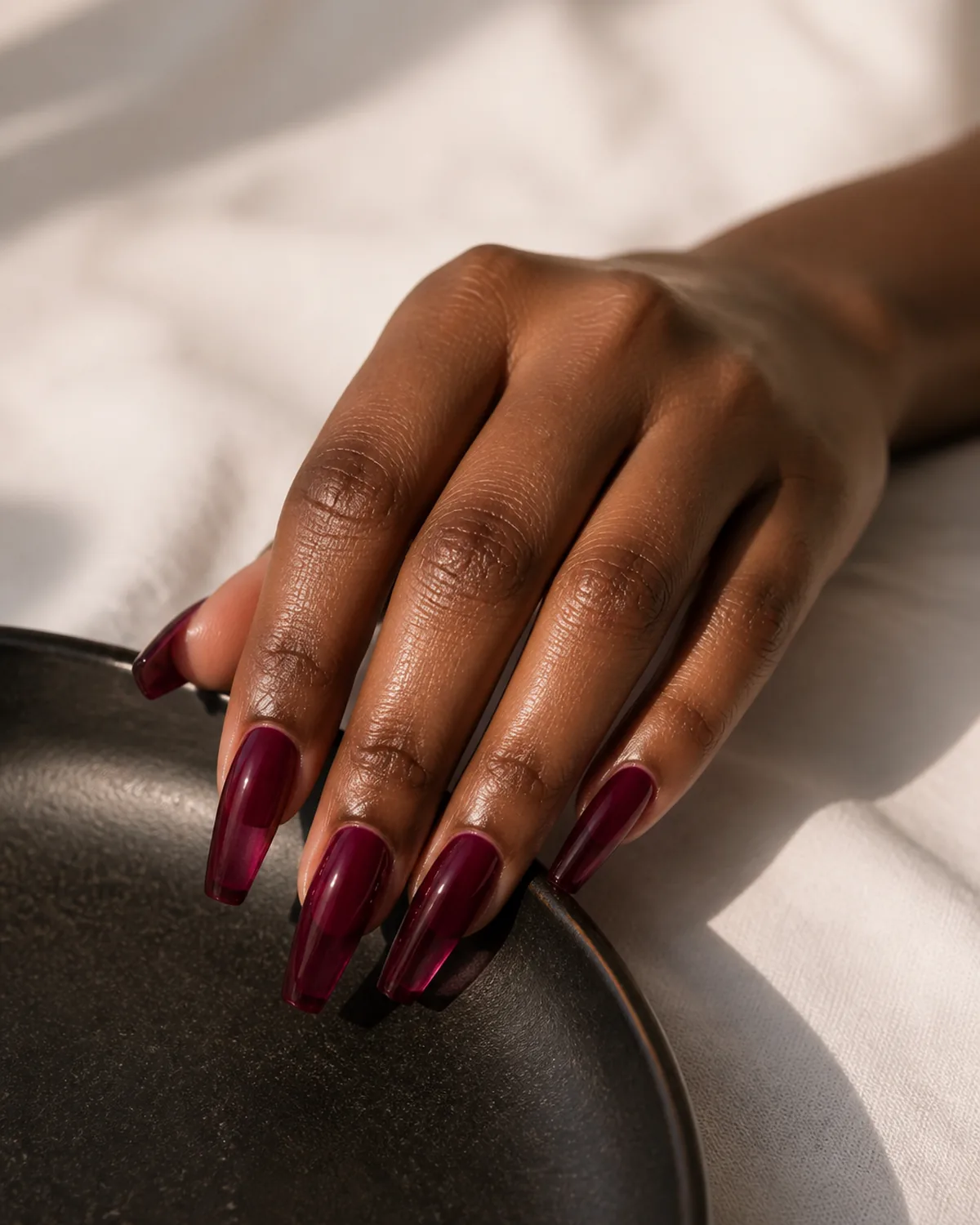 Single hand showing deep blackberry-purple jelly polish on long coffin nails with fingertips resting on the edge of a dark ceramic plate on white linen