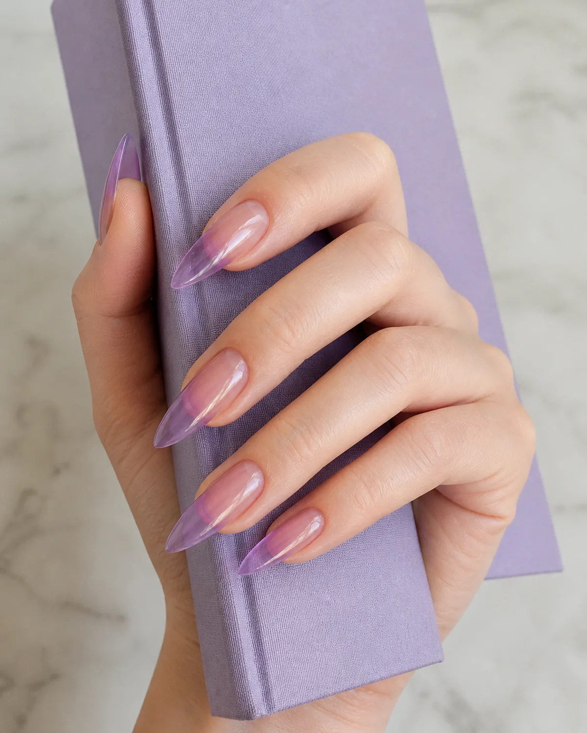 Single hand showing soft purple-lavender jelly polish on pointed stiletto nails with fingers draped along the spine of a lilac hardcover book