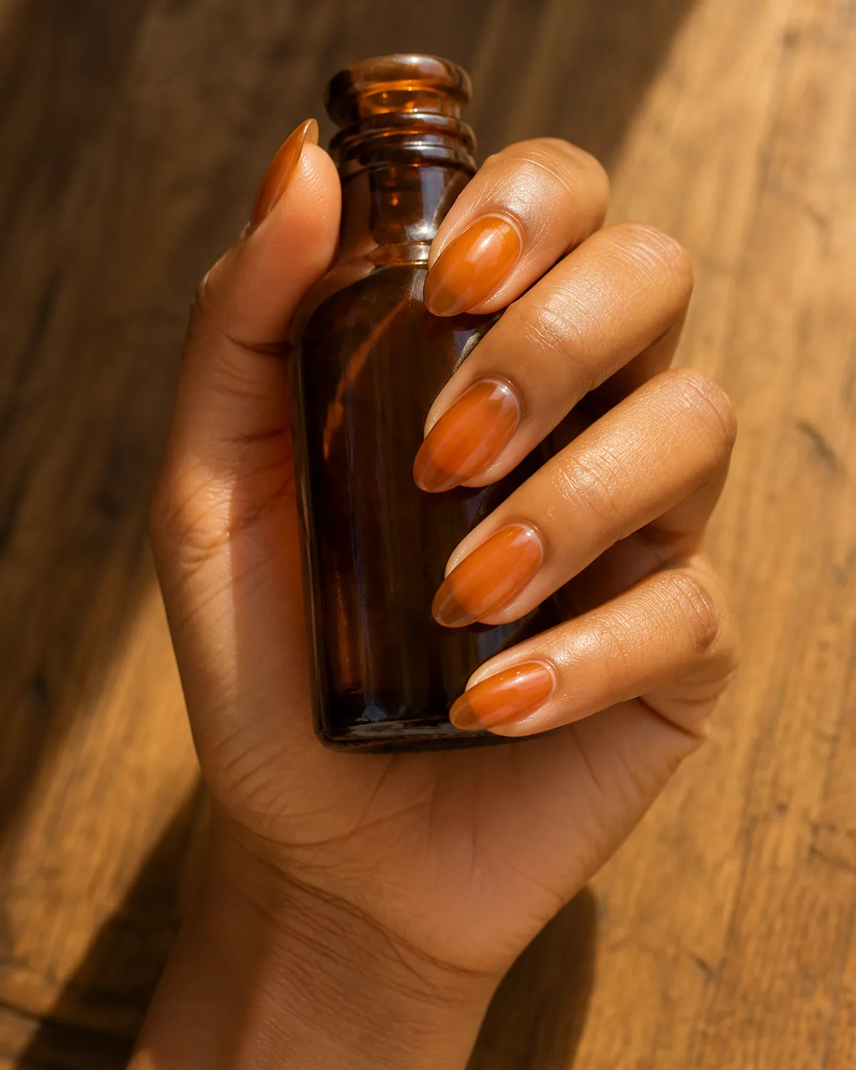 Single hand showing warm golden-brown amber jelly polish on medium almond nails with fingers gently wrapping around the base of an amber glass bottle on warm wood
