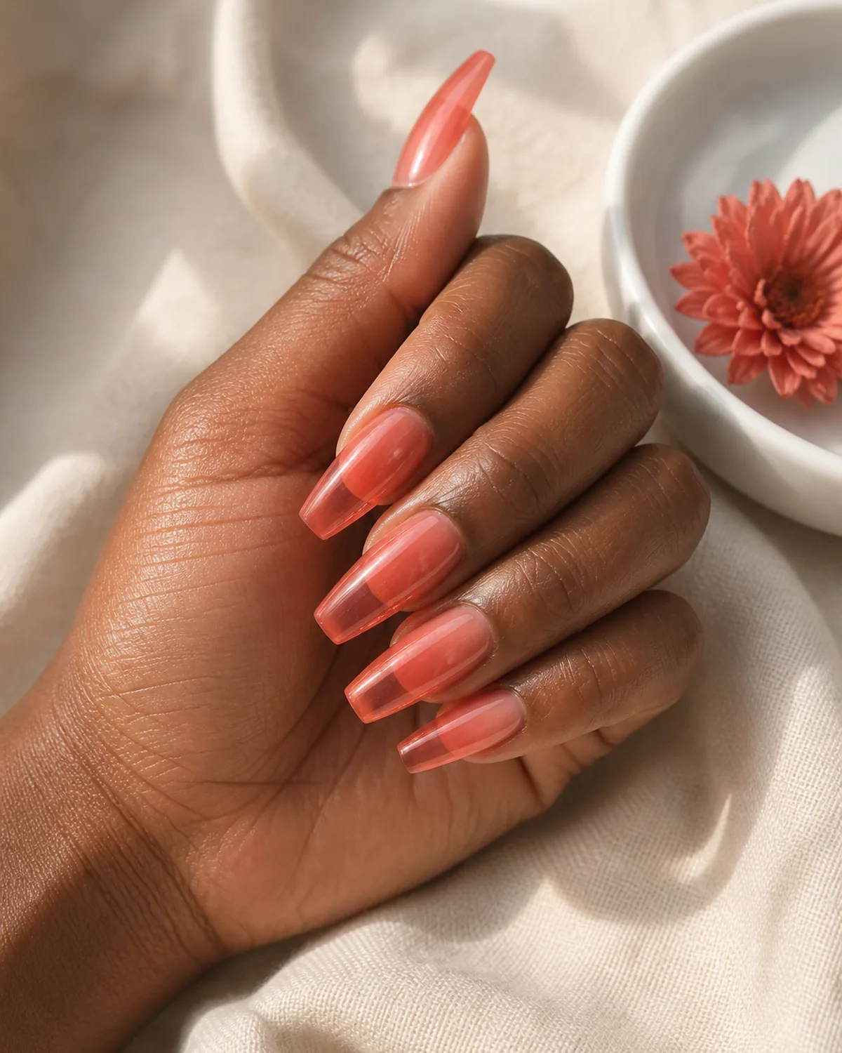 Single hand showing bright warm coral jelly polish on coffin nails resting on its side on cream linen beside a white ceramic bowl with coral dried flower