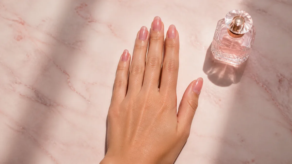 Single hand with sheer rose jelly polish on medium almond nails resting on pale blush marble beside a small glass perfume bottle