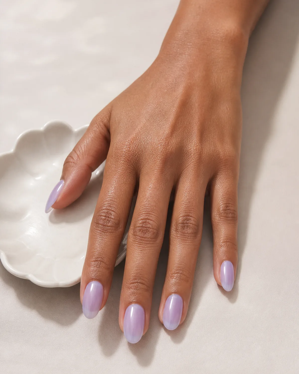 Single hand with sheer lilac jelly-finish polish on medium almond nails beside a small white porcelain dish on cream surface