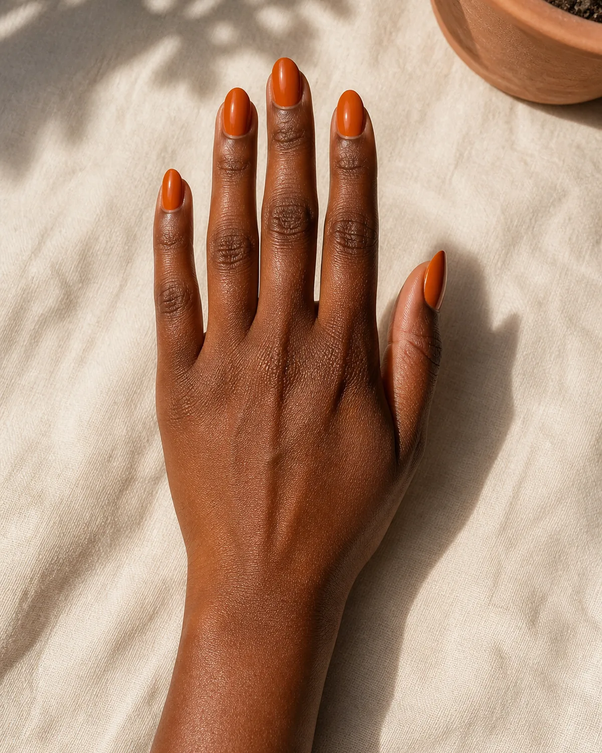 Single hand with rich burnt orange creme polish on medium almond nails resting palm-down on cream linen tablecloth beside small terracotta pot
